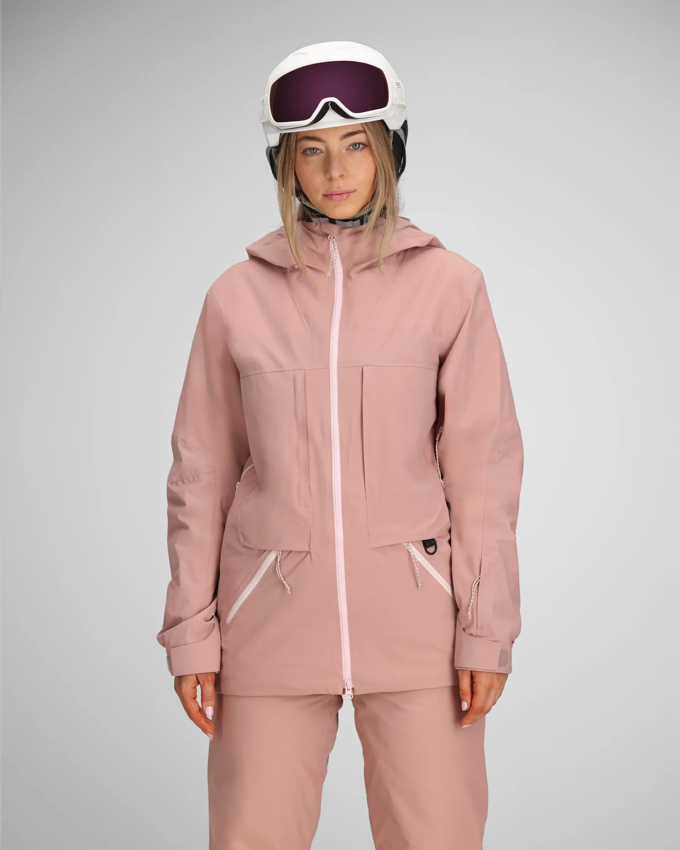A person in a light pink ski outfit and white helmet with goggles stands against a gray background, conveying a sense of calm and preparation.