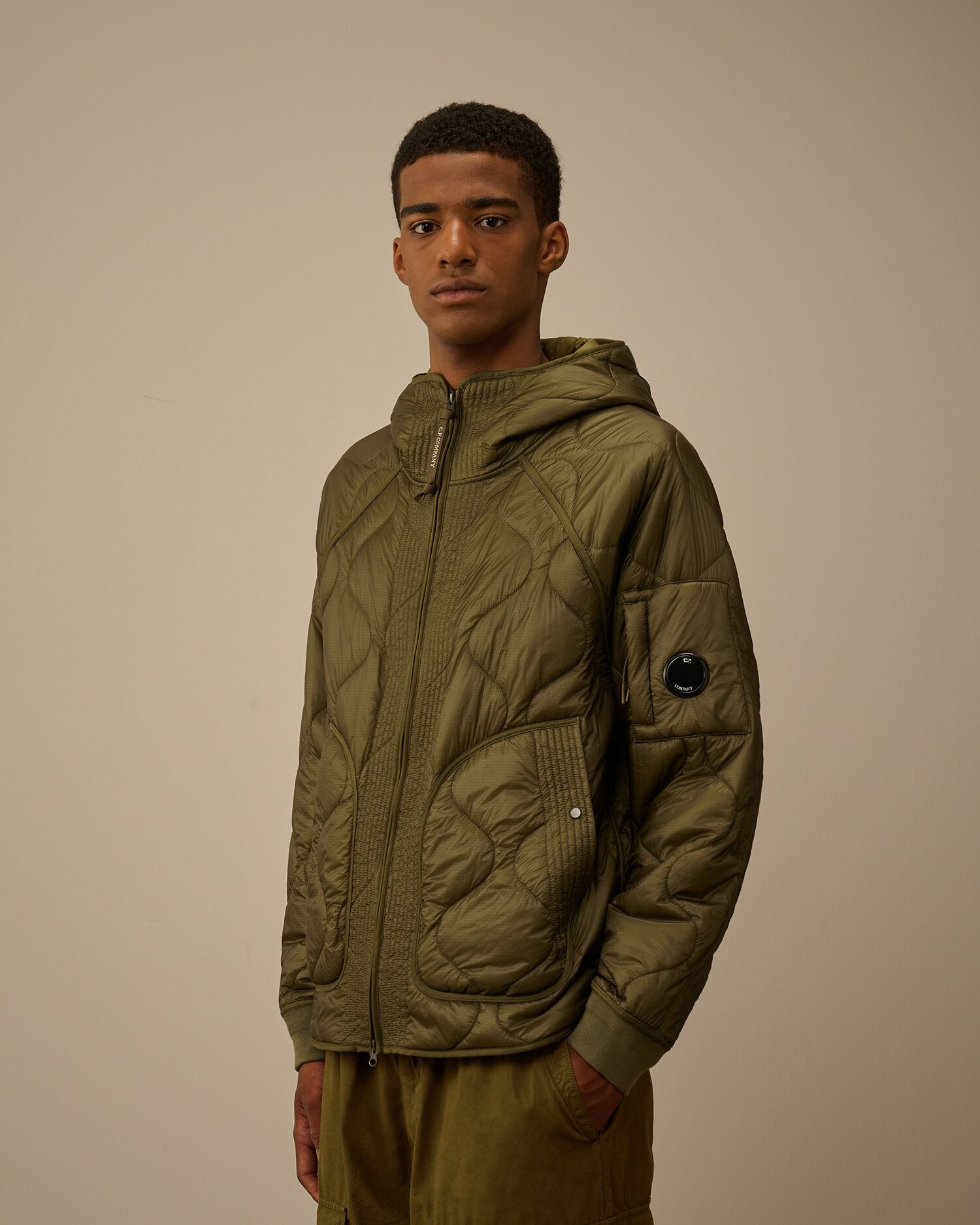 Young man in an olive green quilted jacket with a hood, standing against a neutral background. The mood is calm and casual, with hands in pockets.