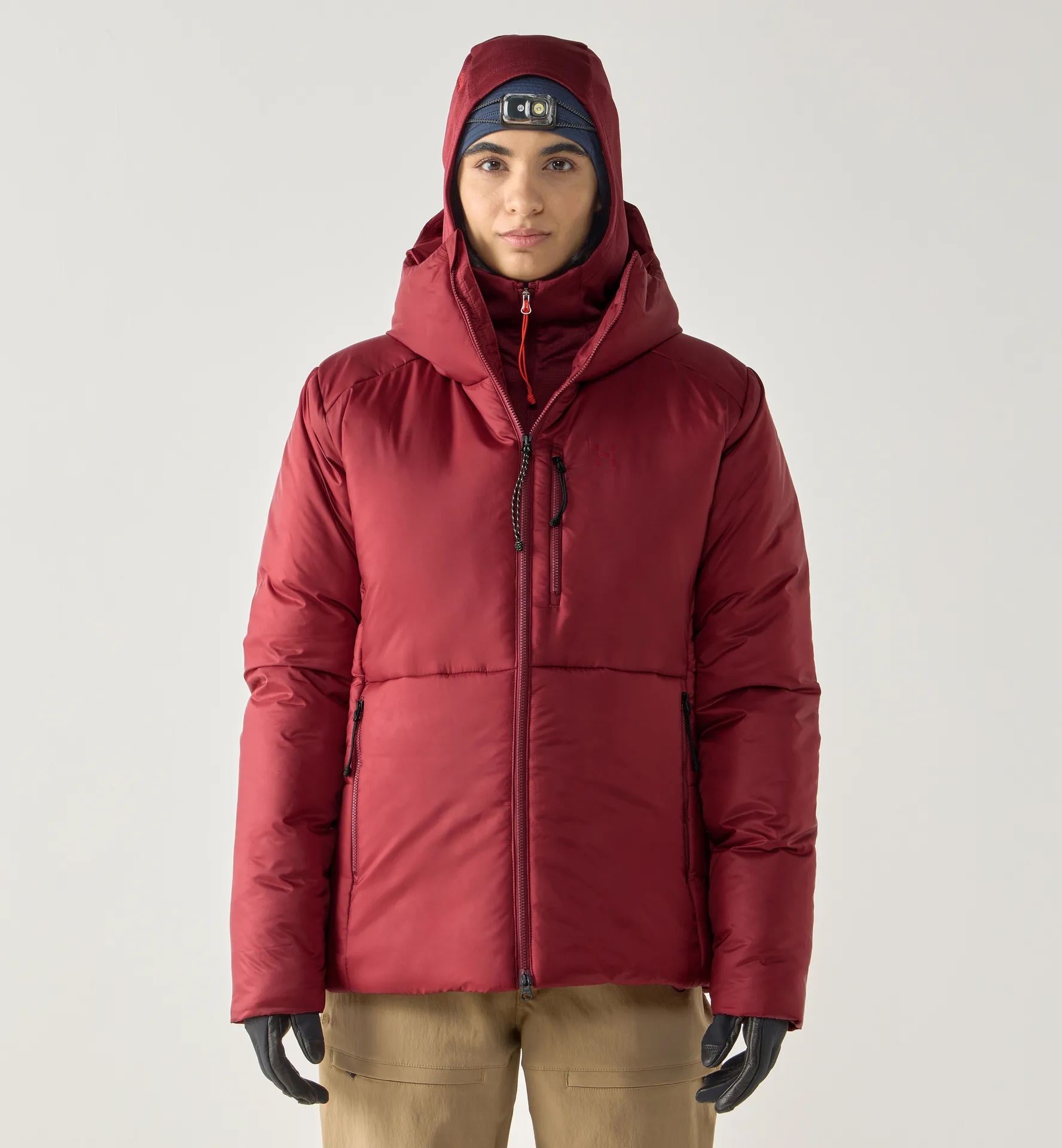 A person in a red puffer jacket with a hood and gloves, wearing a headlamp on a cap. The background is light gray, conveying a sense of warmth and readiness for cold weather.