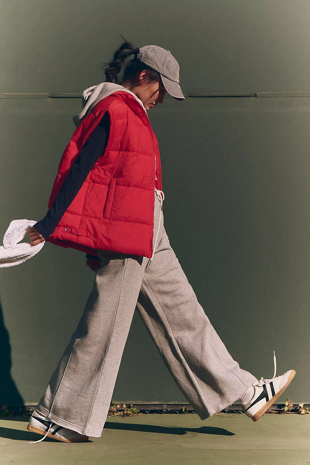 A person in a red puffer vest and gray sweatpants walks casually, wearing a cap. The gesture and outfit suggest a relaxed, sporty vibe against a plain backdrop.