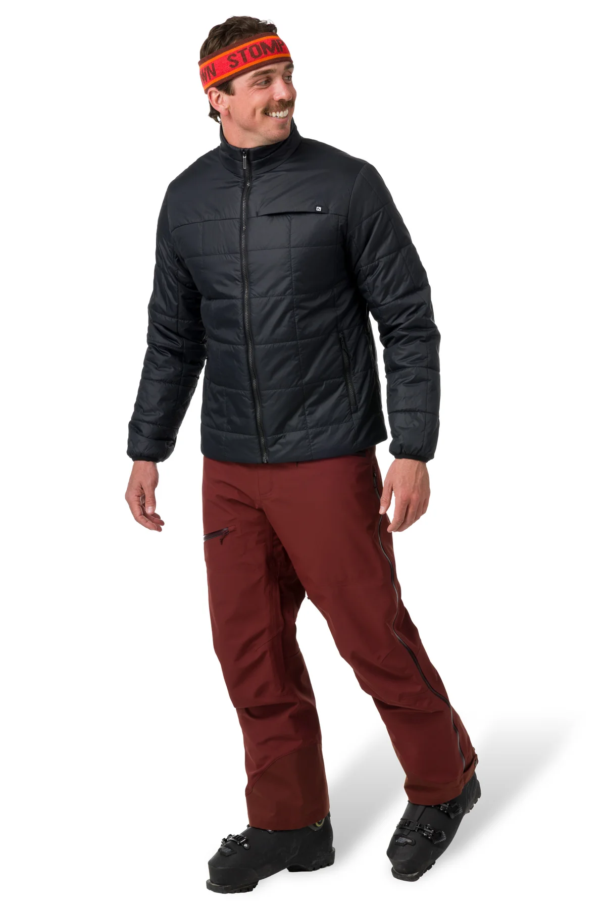 Smiling man wearing a black quilted jacket, maroon ski pants, black boots, and an orange headband. The background is plain white.