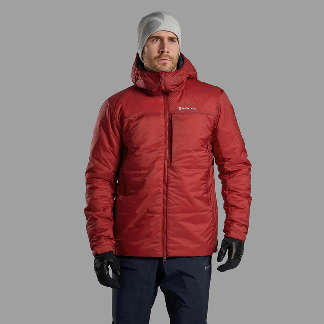 Man in a red insulated jacket with hood looks upward, wearing black gloves, dark pants, and a gray beanie. The background is plain gray.