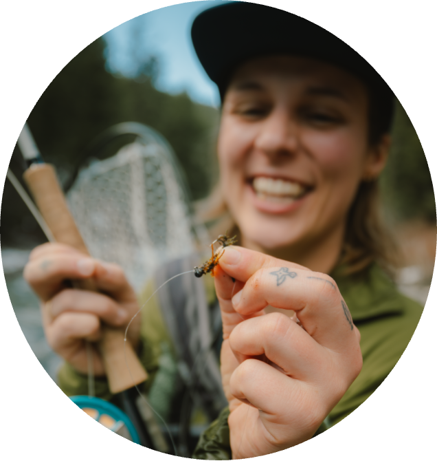 Smiling person holds a fishing rod and a fly fishing lure, showcasing it to the camera. The background features a blurred forest and river.