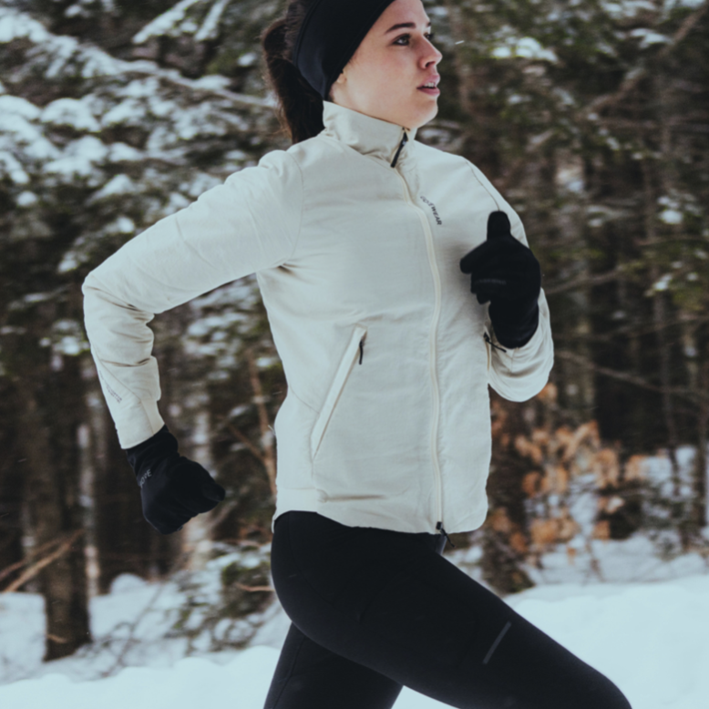 A woman jogs through a snowy forest, wearing a white jacket, black leggings, gloves, and a headband. Her expression is focused and determined.