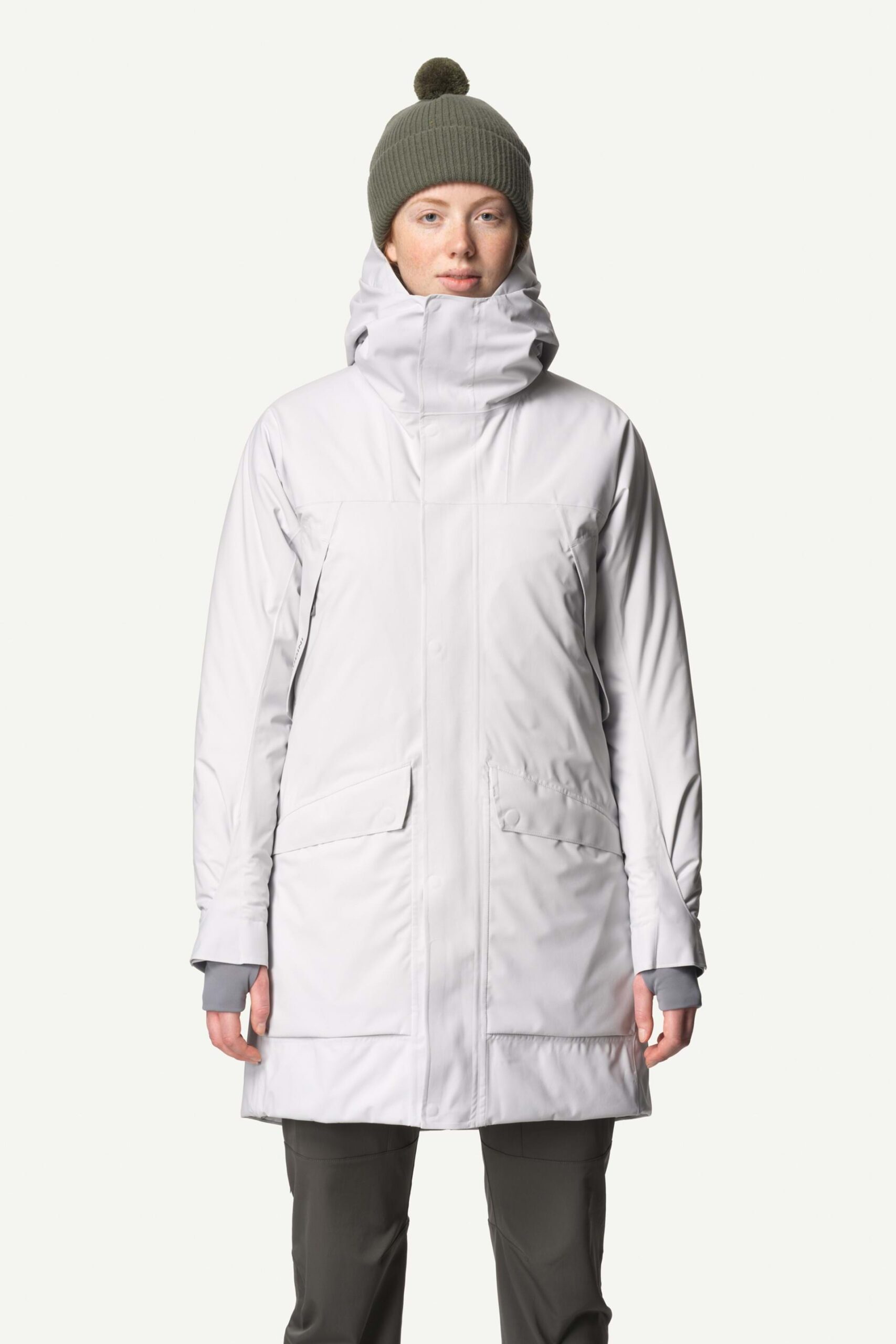 A person models a long, white winter parka with large pockets and a high collar. They wear a green knit beanie with a pom-pom. The mood is practical and cozy.