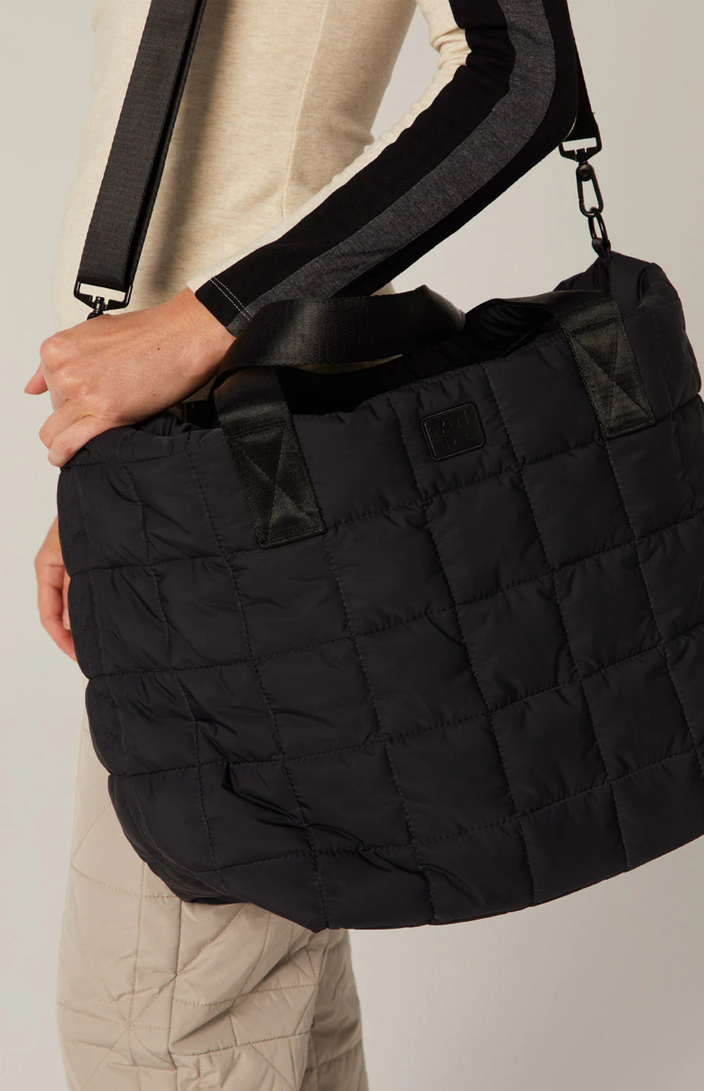 Person holding a large, black quilted tote bag with sturdy straps. They wear a beige top with black sleeves and quilted light pants, creating a cozy, casual look.