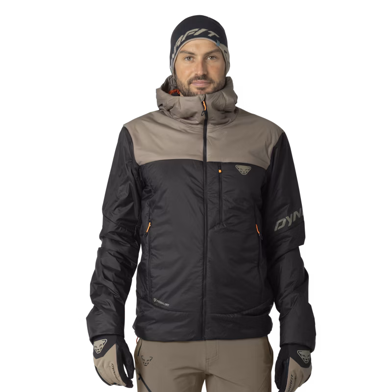 Man wearing a black and gray winter jacket with a high collar and matching gloves, standing against a black background. He has a neutral expression.