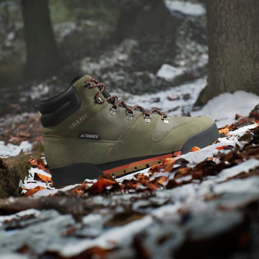 A green hiking boot with red laces sits on snowy, leaf-covered ground in a foggy forest. The scene conveys an adventurous, rugged feel.