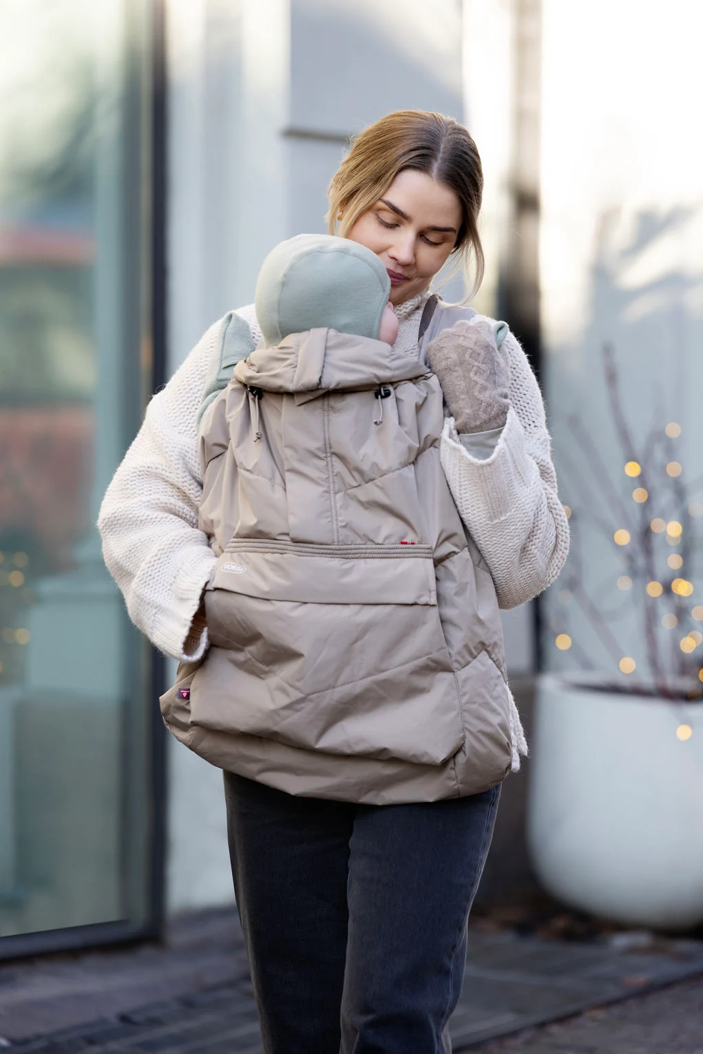 A woman lovingly holds a baby wrapped in a beige carrier, wearing a beanie. She is outside in a casual, cozy setting, exuding warmth and tenderness.